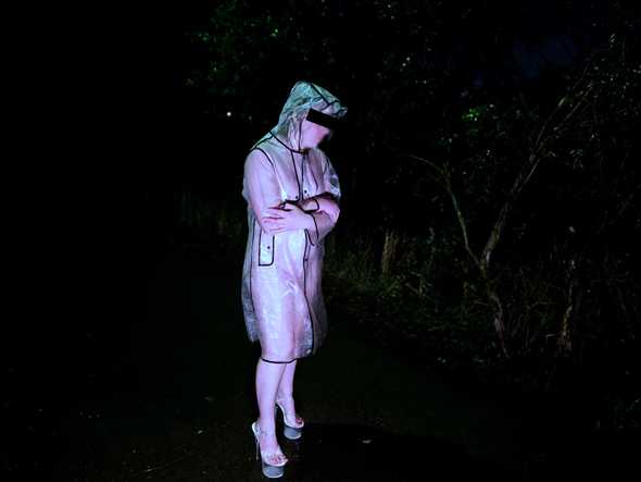 I'm standing alone on a dark rainy night. Shadowy trees are visible in the background. I'm wearing a clear raincoat with nothing on underneath, as well as tall glassy platform heels. I am facing partially towards the camera. My arms are crossed against my chest for warmth. A faint grimace is visible on my face. The photo looks cold.