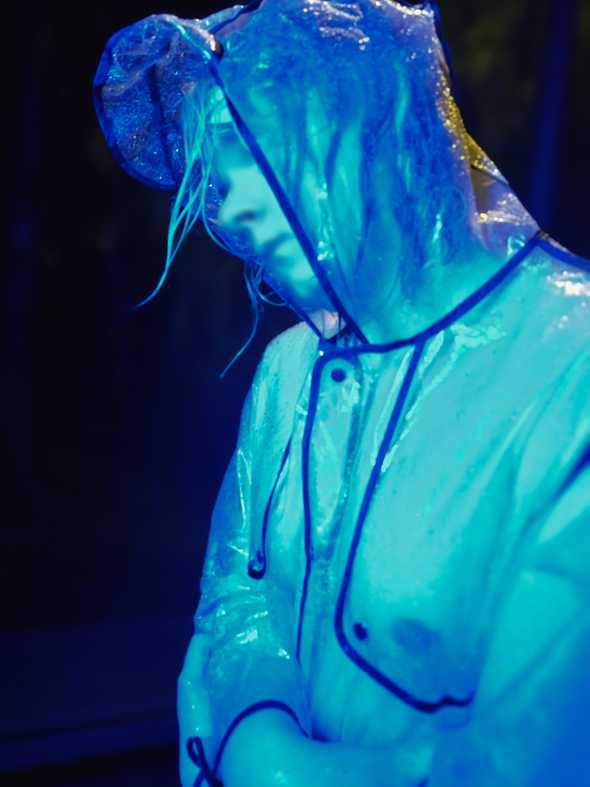 A blurred close-up of my torso & face. I am hugging myself to try to stay warm. My nipples are visible through the transparent raincoat. I have a melancholic expression.